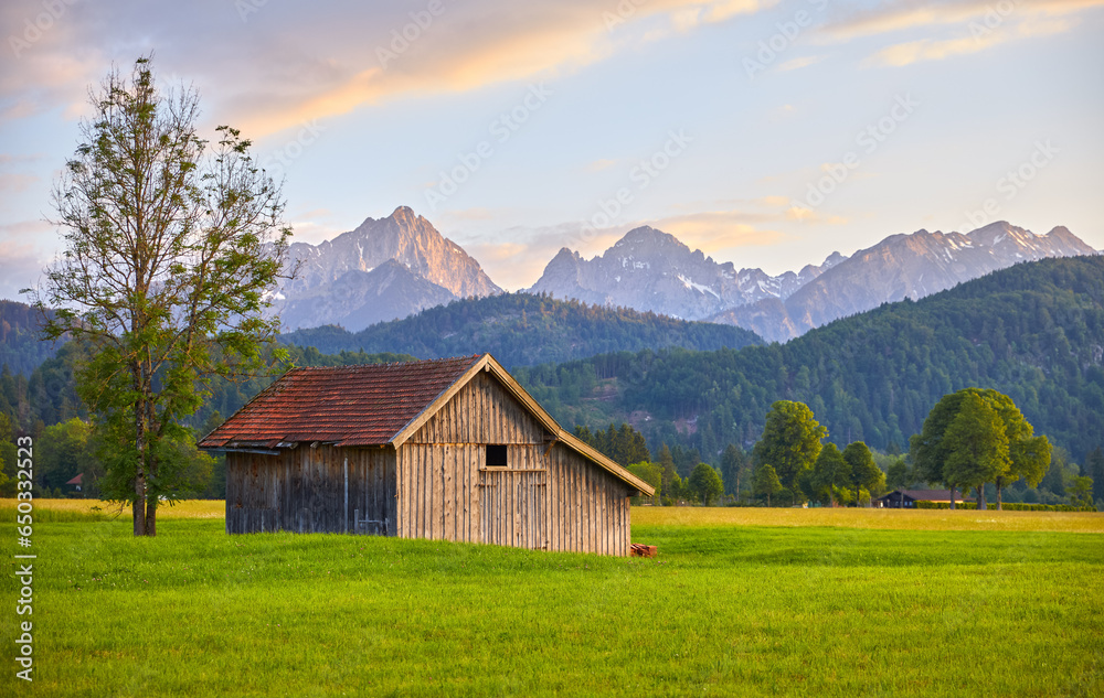 Wooden barn on meadow in Bavaria, Germany. Panorama of green field with grass and picturesque german Alps mountains during sunset