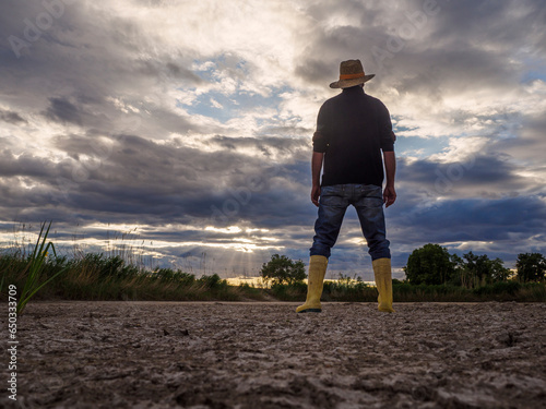 With rubber boots for the dry field. The farmer goes with his rubber boots and his shovel to a barren field. View from behind. Drought concept.