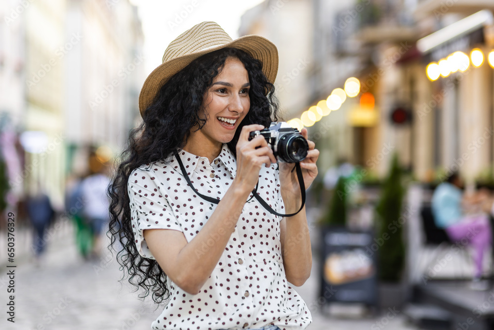 Young beautiful hispanic woman with curly hair walking in the evening city with a camera, female tourist on a trip exploring historical landmarks in the city.