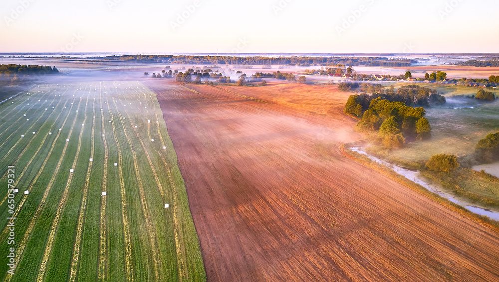 Warm sunny September morning. Autumn beautiful fog landscape. Small river curves in trees. Bright sunny misty riverside. Scenic view of rural sunrise. Village in Agriculture fields.