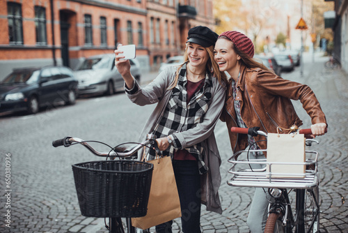 Fototapeta Naklejka Na Ścianę i Meble -  Two young female friends riding bikes while shopping in the city