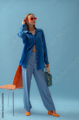 Fashionable young woman wearing trendy orange sunglasses, blue linen shirt, trousers, block heel shoes, holding bag, posing on blue background. Full-lenght studio portrait. Copy, empty space for text