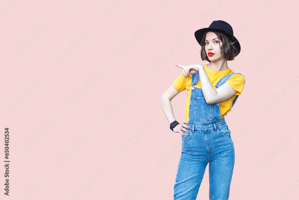 Portrait of surprised amazed hipster woman in blue denim overalls, yellow T-shirt and black hat, pointing at copy space for advertisement. Indoor studio shot isolated on light pink background. Portrait of surprised amazed hipster woman in blue denim overalls, yellow T-shirt and black hat, pointing at copy space for advertisement. Indoor studio shot isolated on light pink background.