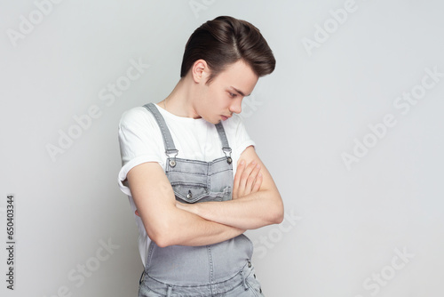 Portrait of sad unhappy young brunette man standing with folded hands, frowns face upset with bad news he received, wearing denim overalls. Indoor studio shot isolated on gray background.