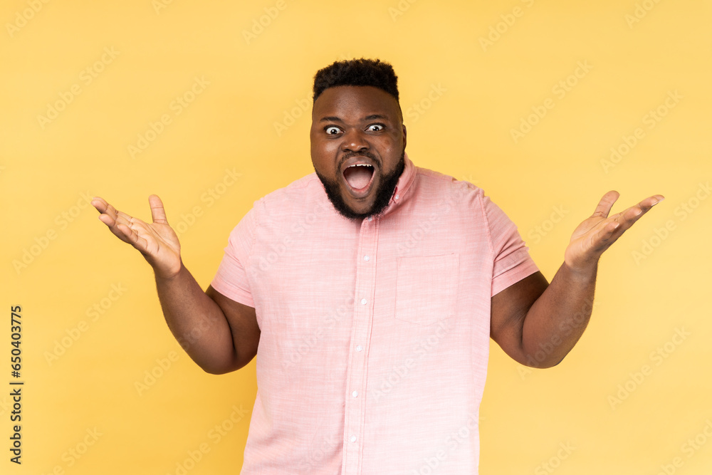 What a surprise. Portrait of man wearing pink shirt standing with wide open mouth and raising hands in amazement, shocked by unbelievable success. Indoor studio shot isolated on yellow background.
