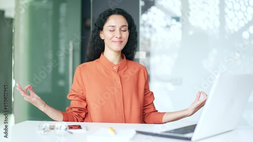 Young female employee is meditating with her eyes closed sitting at a workplace in office. Businesswoman took a break from work to relieve stress. Brunette woman rests, relaxes, feels peace of mind photo