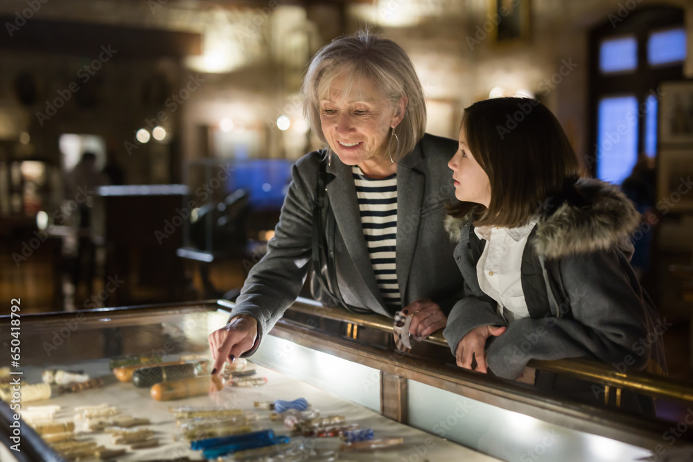 Friendly aged female tutor helping preteen girl exploring antique showpieces in local history museum..