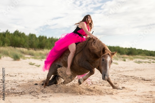 Female rider wearing a pink dress galloping along a beach on a brown horse.