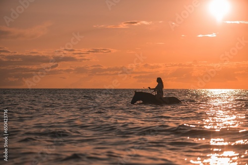 Female riding a horse through a shallow body of water at sunset