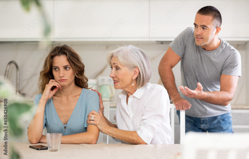 Fototapeta premium Young girl is sitting at table in kitchen quarreling with her mother-in-law and husband