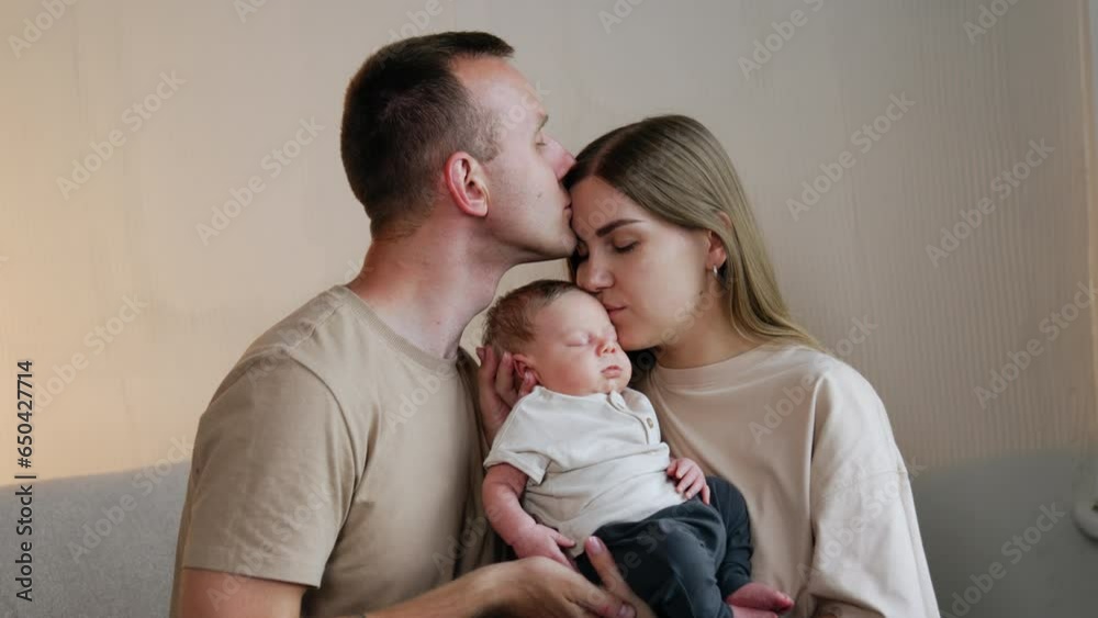 Long-haired woman kisses her sleeping newborn on the head. Loving man kisses his wife on the forehead.
