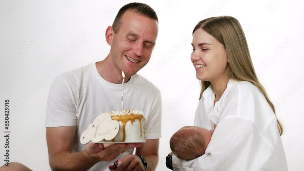 Smiling Caucasian couple with baby blowing a candle on the cake. Family celebrating first month of their infant. White backdrop. Smiling Caucasian couple with baby blowing a candle on the cake. Family celebrating first month of their infant. White backdrop.