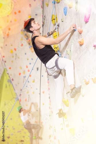 Well-equipped, positive young man training on stone wall, artificial mountain with rocks, climbing up in bouldering park indoors
