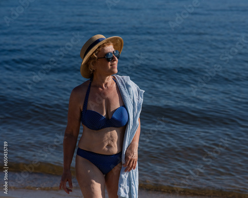 An old woman in a straw hat, sunglasses and a swimsuit is resting on the beach. 