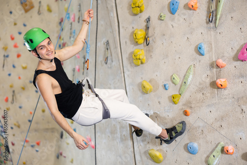 Sporty young guy in safety gear doing a difficult wall climb in climbing gym