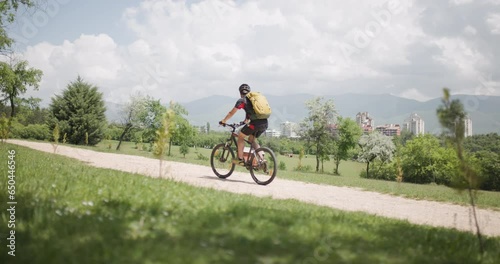 Male mountain biker fully geared riding his mountainbike uphill on a pebbles dirt road near a green field. Exploring dirt road in a scenic meadow. Recorded on cinema camera. photo