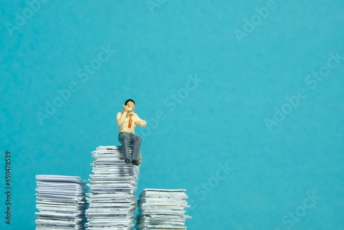 Miniature tiny people toy figure photography. Call list concept. A businessman sitting above pile of document while make a phone call. Isolated on blue background