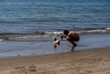 Hombre en la playa tomándole una foto a un perro maltés que descansa en la arena mientras aprecia la inmensidad del mar.