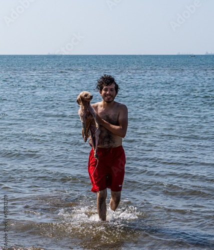 Hombre joven en traje de baño, feliz cargando a su perro en la orilla de la playa, disfrutando de una hermosa tarde calurosa en compañía de su mejor amigo. photo