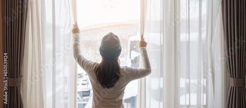 young woman in sweater looking through the window in winter season, happy female rising arms and stretching after waking at apartment or home in the morning. Lifestyle and Relaxing concepts