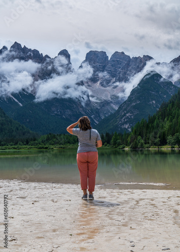 A middle-aged woman travels the world. Stands near the lake against the backdrop of the majestic mountains of the Dolomites, South Tyrol. Italy. Back view. © Alexander