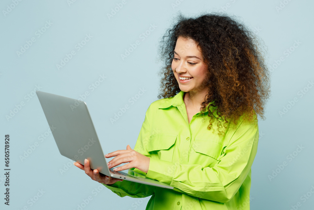 Side view smiling young IT latin woman she wear green shirt casual clothes hold use work on laptop pc computer isolated on plain pastel light blue cyan background studio portrait. Lifestyle concept. Side view smiling young IT latin woman she wear green shirt casual clothes hold use work on laptop pc computer isolated on plain pastel light blue cyan background studio portrait. Lifestyle concept.