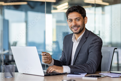 Portrait of young hispanic businessman inside office at workplace, man holding bank credit card in hands, using laptop for online shopping and money transfer, employee smiling and looking at camera.