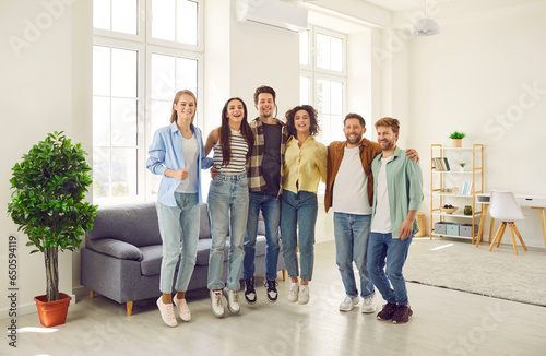 Portrait of a group of young happy smiling friends students or colleagues in casual clothes standing together in the living room at home, hugging and looking cheerful at the camera.