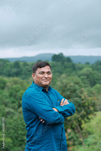 Happy mature Indian asian man with arms crossed standing at nature , Old mustache and brown haired male smiling wearing blue shirt. "selective focus" " shallow depth of field" or " blur". 