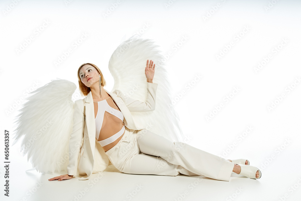 full length of charming woman in costume of heavenly angel with wings sitting on white backdrop