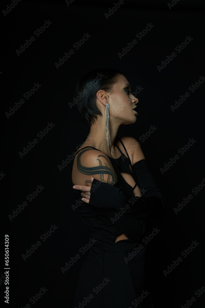 side view of enchanting tattooed woman in black halloween dress and shiny earring on black backdrop side view of enchanting tattooed woman in black halloween dress and shiny earring on black backdrop