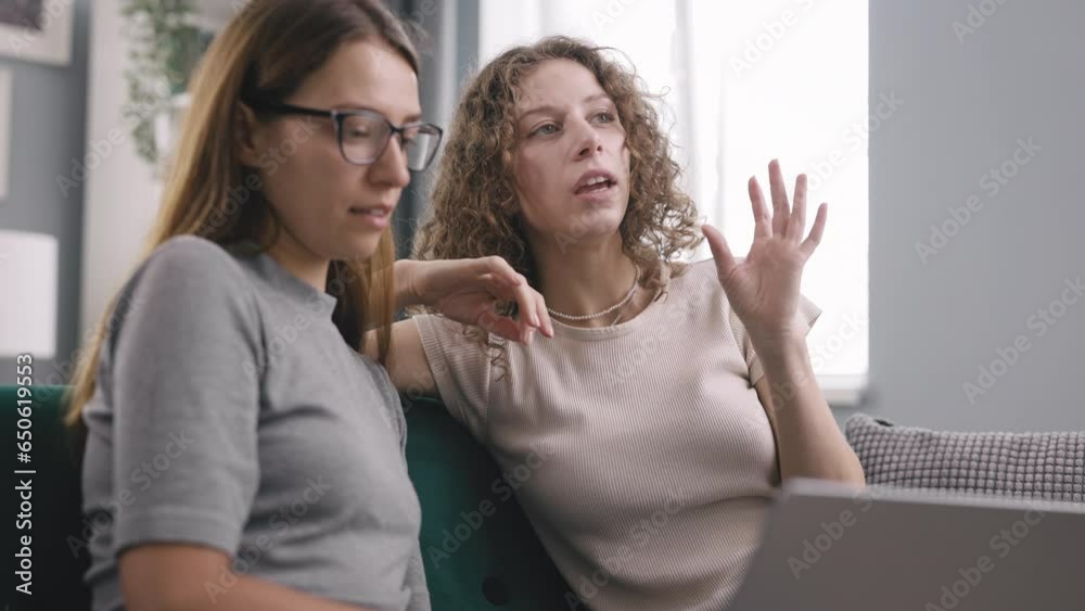 Two friends browsing on laptop on couch, curly woman explain friend in glasses what exactly need and together search for this on Internet Two friends browsing on laptop on couch, curly woman explain friend in glasses what exactly need and together search for this on Internet