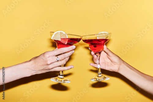 Close up of two female hands holding red cocktails against vibrant yellow background, copy space