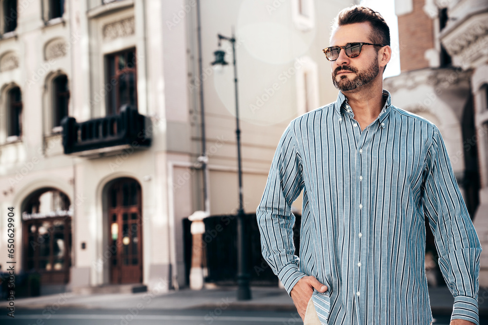 Obraz premium Portrait of handsome confident stylish hipster lambersexual model. Sexy modern man dressed in blue shirt and trousers. Fashion male posing on street background in Europe city at sunset. In sunglasses