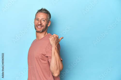 Middle-aged caucasian man on blue backdrop points with thumb finger away, laughing and carefree.