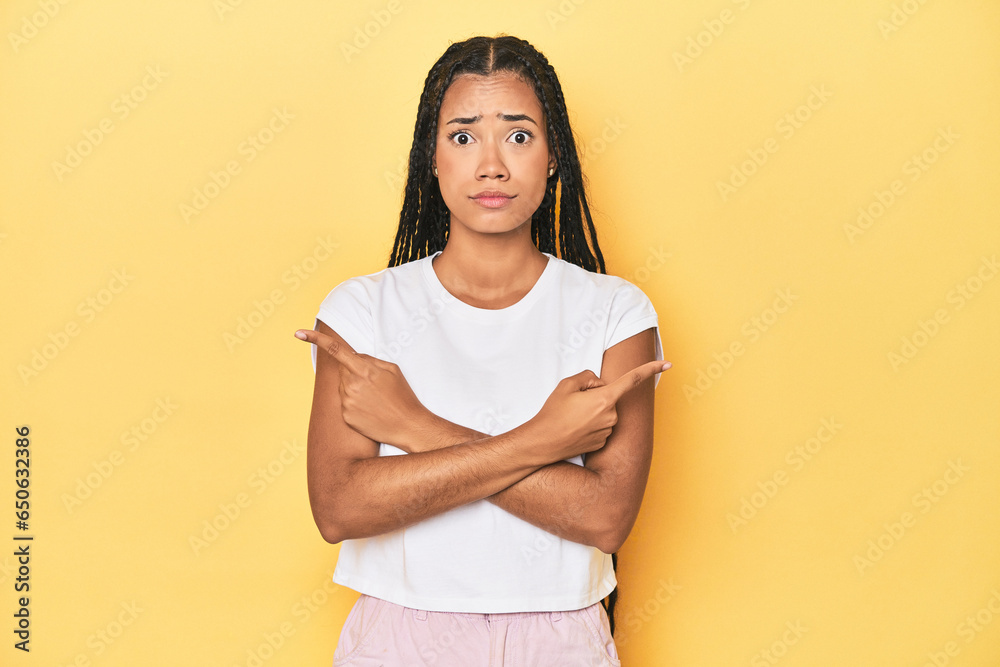 Young Indonesian woman on yellow studio backdrop points sideways, is trying to choose between two options.