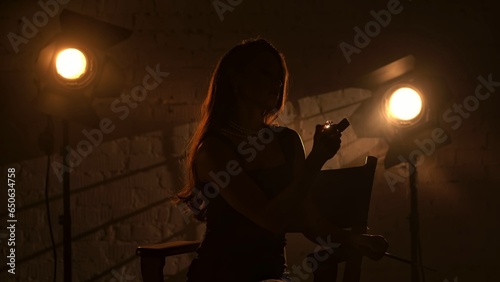 Tableau sur toile Woman in black dress and pearl necklace sitting on a director's chair, holding cigarette holder and tries to light up the lighter