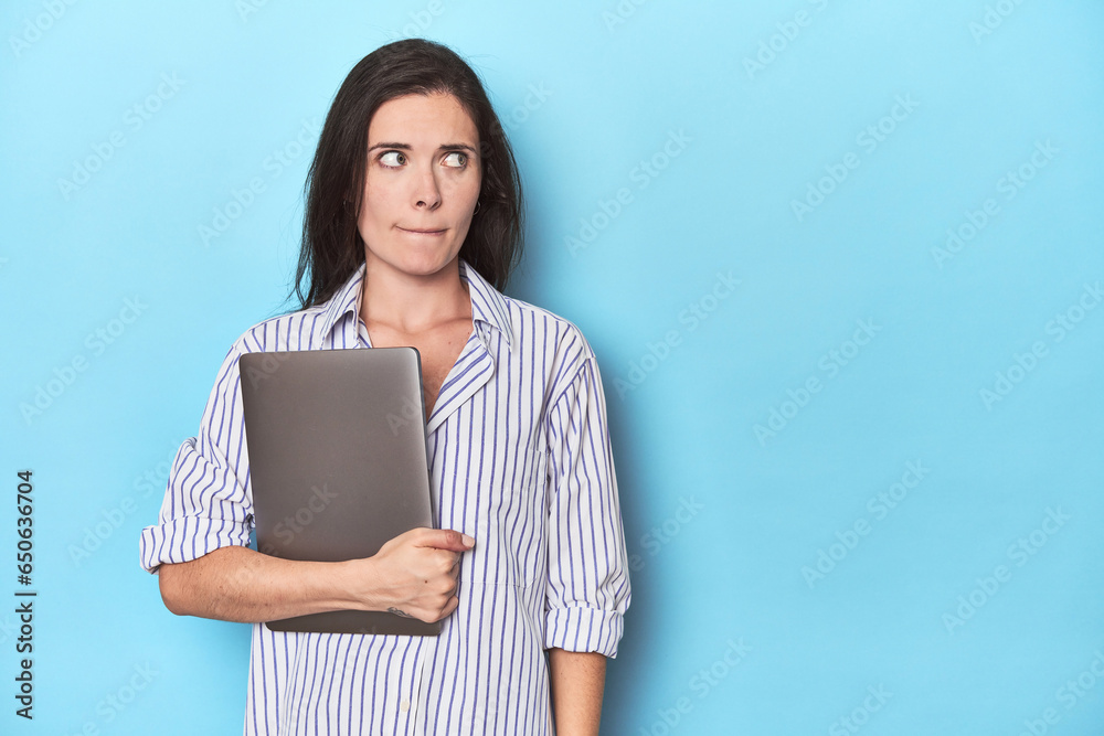 Young businesswoman holding laptop on blue confused, feels doubtful and unsure. Young businesswoman holding laptop on blue confused, feels doubtful and unsure.