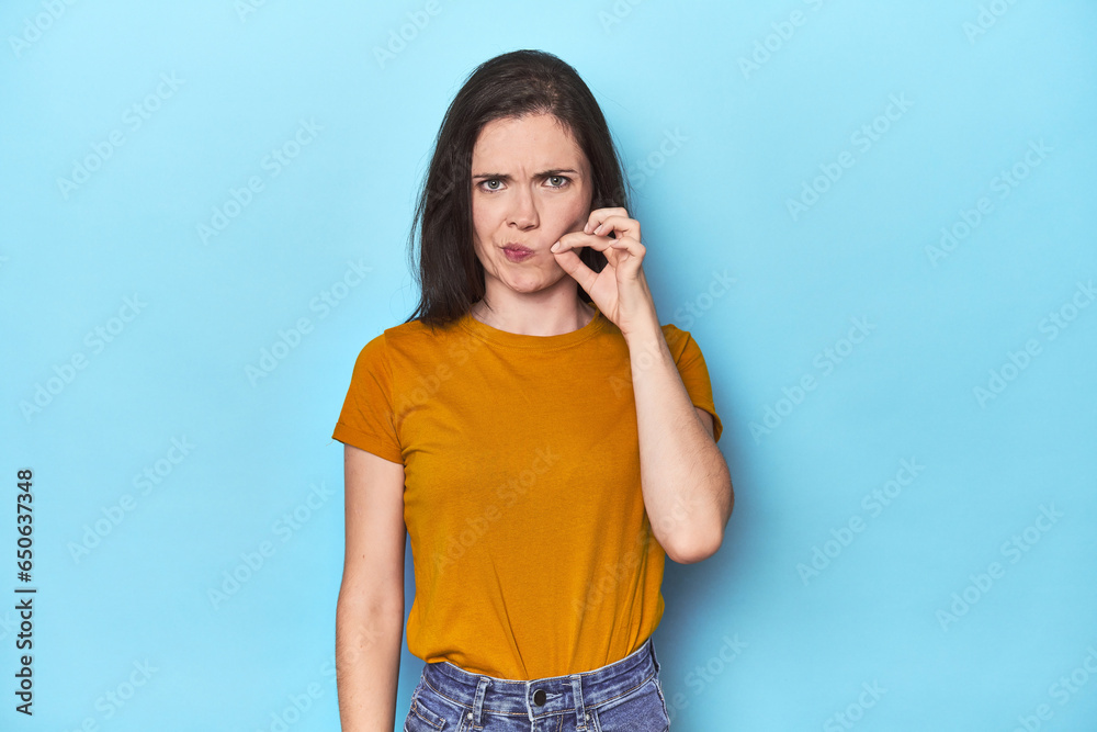 Young caucasian woman on blue backdrop with fingers on lips keeping a secret. Young caucasian woman on blue backdrop with fingers on lips keeping a secret.