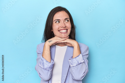 Woman in blue blazer on blue background keeps hands under chin, is looking happily aside.