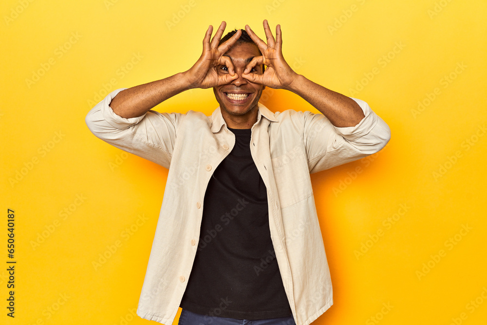 African American man with shirt, yellow studio, showing okay sign over eyes African American man with shirt, yellow studio, showing okay sign over eyes