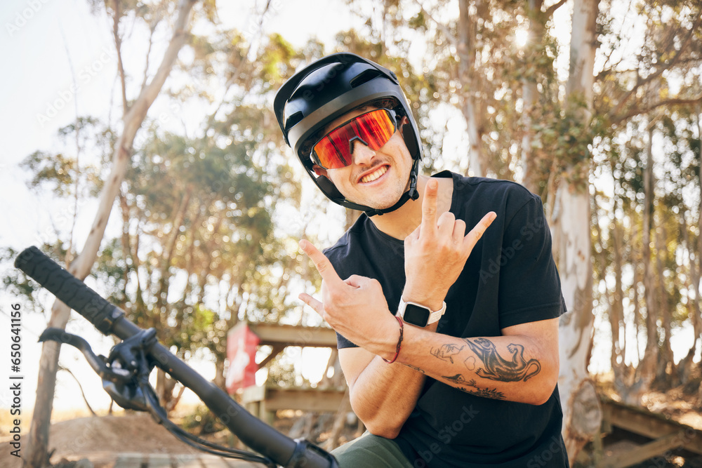 Obraz premium Rock sign, bike and man with helmet in forest on off road cycling workout with happiness and glasses. Portrait, person and smile with hand gestures on bicycle for exercise, challenge and competition