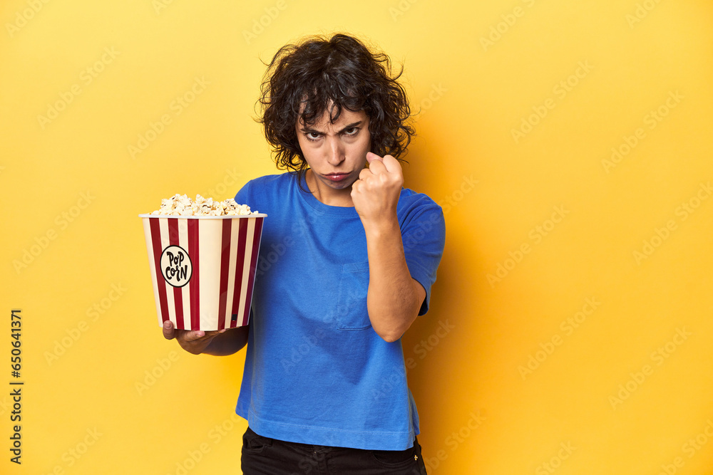 Curly-haired woman with popcorn for movie, studio showing fist to camera, aggressive facial expression. Curly-haired woman with popcorn for movie, studio showing fist to camera, aggressive facial expression.