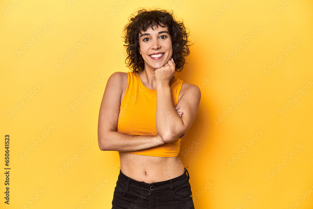 Curly-haired Caucasian woman in yellow top smiling happy and confident, touching chin with hand.