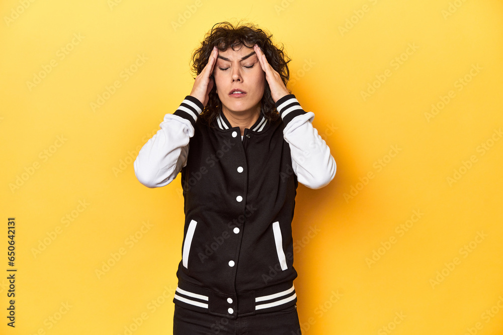 Curly-haired Caucasian woman in baseball jacket touching temples and having headache. Curly-haired Caucasian woman in baseball jacket touching temples and having headache.