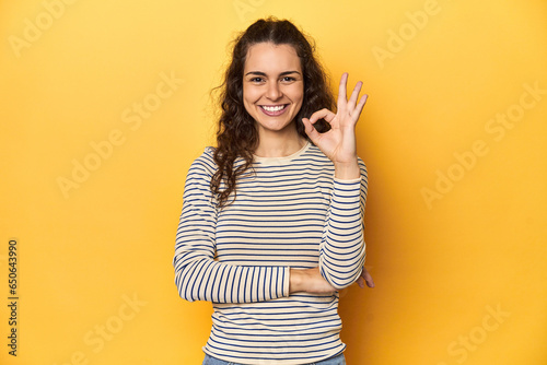 Young Caucasian woman, yellow studio background, winks an eye and holds an okay gesture with hand.