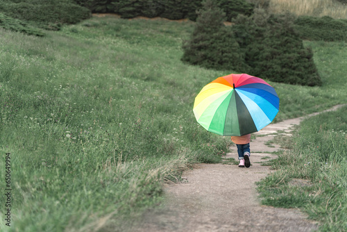 Child with a rainbow umbrella. Back view. A girl walks against a green background of nature. Childhood concept