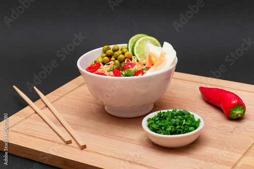 Beautiful Noodle Dish with Green Pea, Eggs, Red Hot Pepper, Greens and Lime with Chopsticks on Bamboo Cutting Board. Instant Noodles