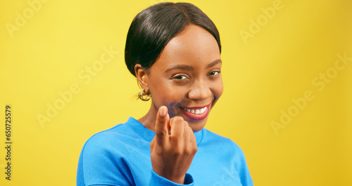 Flirty young woman beckons with finger 'come here', yellow studio background
