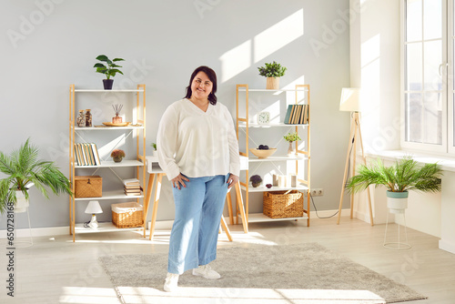 Full lenght portrait of a fat plus size brunette woman standing at home in the living room and smiling. Plump overweight cheerful girl looking positively at the camera. Body positive concept.